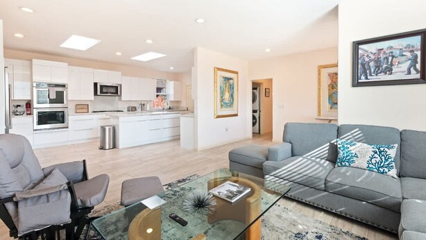 A modern living room with a glass coffee table, grey sofa, and armchairs. A kitchen with white cabinets and appliances is visible in the background. - Spacious Single Level Home In Poway (Poway)