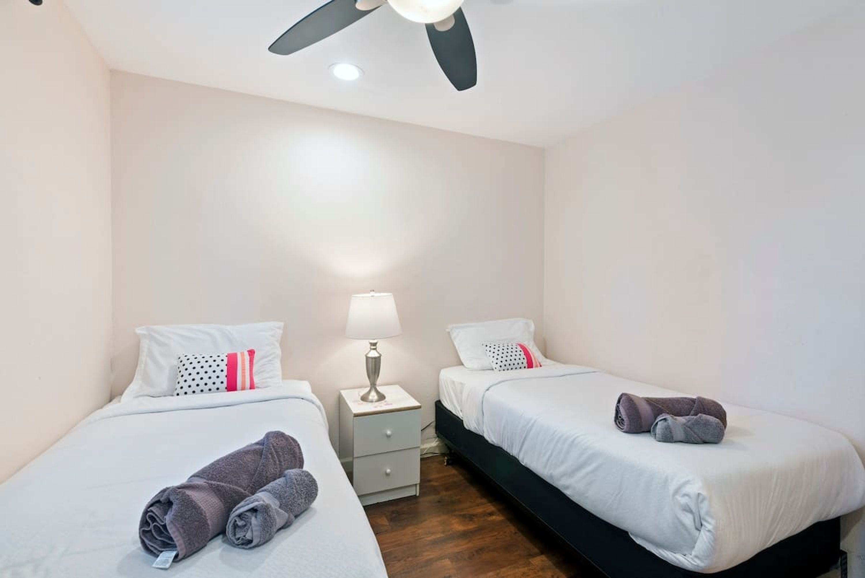 A hotel room with two single beds, a nightstand with a lamp, and a ceiling fan.