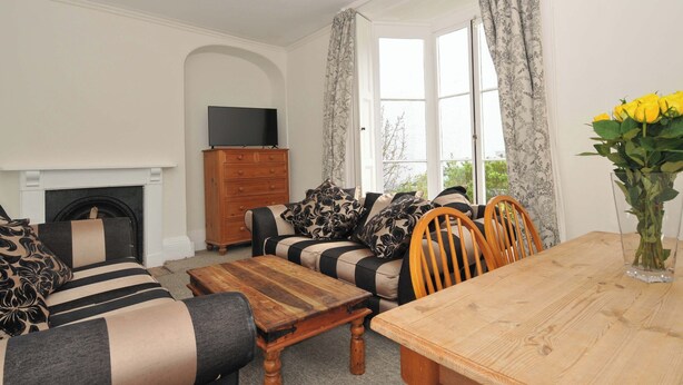 A living room with a sofa, a wooden table, a TV on a dresser, and a fireplace. - Violet's Rest (Exeter)