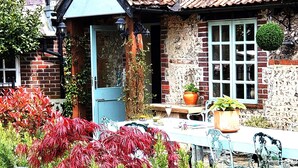 A rustic house with a tiled roof, a brick exterior, and a front door. There is a small table with a potted plant and a chair outside.