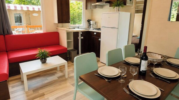 Un espace de vie compact comprenant un canapé rouge, une table à manger pour quatre personnes, un petit coin cuisine et une vue sur un bâtiment à l’extérieur.