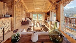 Interior acogedor de una cabaña con paredes y techos de madera, un sofá, una mesa de comedor y vistas a las montañas a través de amplias ventanas.