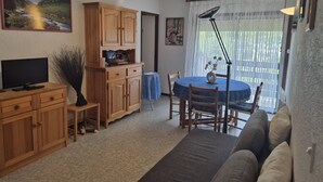 A living room with a TV, wooden cabinets, a dining table with chairs, a sofa, and a large window with curtains. - 2-Bedroom Apartment with Balcony and Parking in Cauterets (Cauterets)