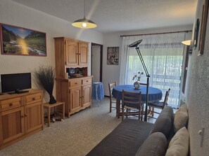 A living room with a TV, wooden cabinets, a dining table with chairs, a sofa, and a large window with curtains. - 2-Bedroom Apartment with Balcony and Parking in Cauterets (Cauterets)