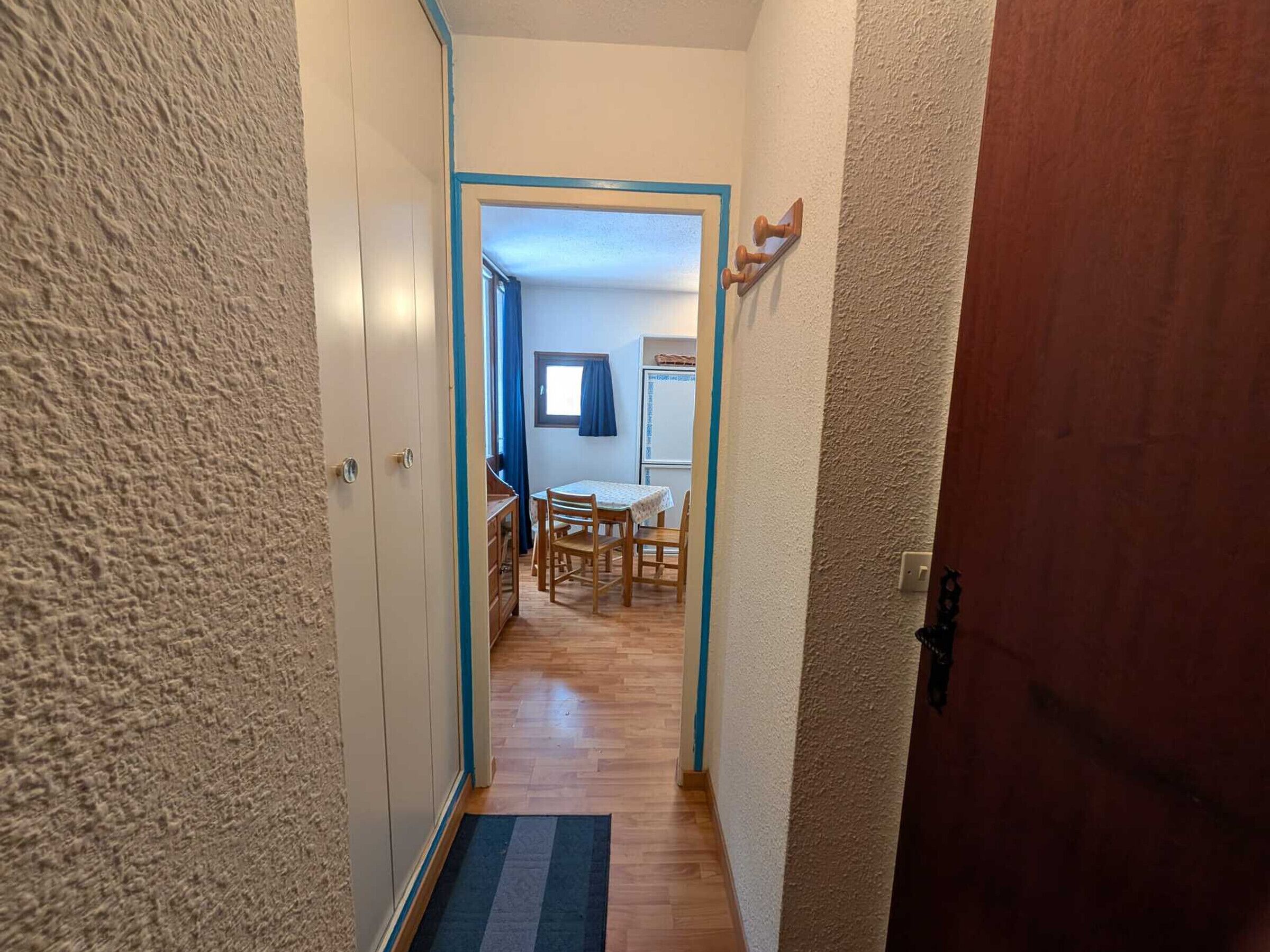A narrow hallway with a wooden floor leading to a dining area with a table and chairs.