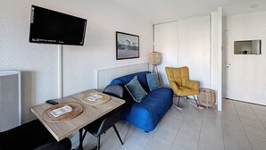 Ein Wohnzimmer mit einer Couch, einem Tisch mit Platzsets, einem an der Wand montierten Fernseher, einem gerahmten Bild und einer Tür.