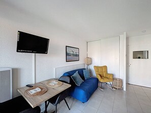 Ein Wohnzimmer mit einer Couch, einem Tisch mit Platzsets, einem an der Wand montierten Fernseher, einem gerahmten Bild und einer Tür.