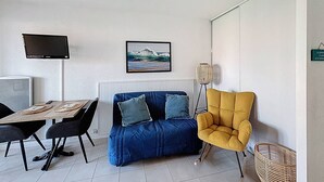 Ein modernes Zimmer mit einer Couch, einem gelben Sessel, einem Esstisch mit Stühlen, einem Fernseher und einem gerahmten Bild an der Wand.