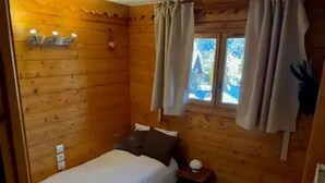 Een houten kamer met een bed, een klein nachtkastje, een raam met gordijnen en een wandlamp.