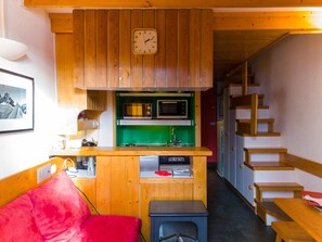 Eine kompakte Küche mit Holzschränken, einer Mikrowelle und einem Spülbecken. Ein rotes Sofa und ein kleiner Tisch mit Heizung sind ebenfalls vorhanden. Eine Treppe ist rechts sichtbar.