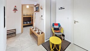 Una habitación con un banco de madera, un taburete amarillo, una silla con cojín estampado y una puerta con un letrero.