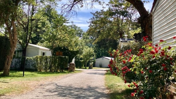 Une allée de gravier mène à une maison blanche dotée d’un porche abrité, le tout entouré d’une végétation luxuriante et d’arbustes en fleurs.