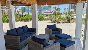 Een overdekt terras met rieten meubels en blauwe kussens, met uitzicht op een strand met palmbomen en campers.