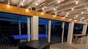 Een buitenterras met een tafel en stoelen, een zwembad op de achtergrond en sfeerverlichting aan het plafond.