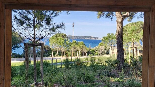 Une vue à travers un cadre en bois sur un jardin agrémenté d’une pergola, d’arbres et, au fond, d’une étendue d’eau.
