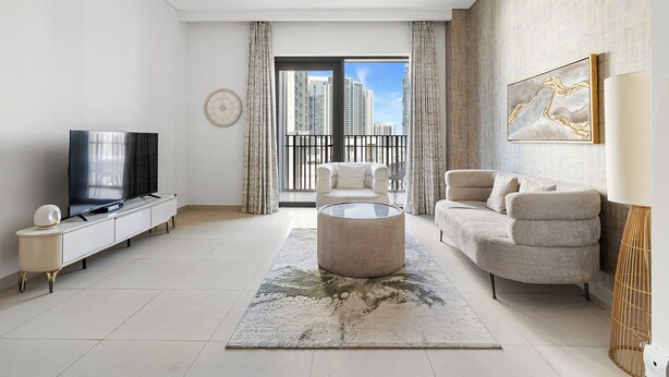 A modern living room with a flat-screen TV, a round coffee table, a sofa, and a large window offering a city view. - 1BR Creek Beach Access (Dubai)
