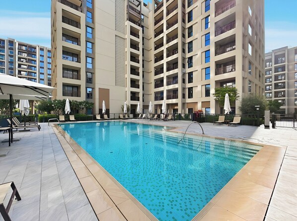A swimming pool surrounded by lounge chairs and umbrellas, with a high-rise building in the background. - 1BR Creek Beach Access (Dubai)