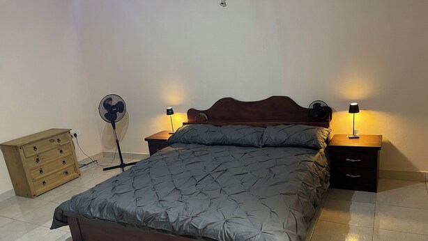 Une chambre à coucher avec un lit, deux tables de chevet avec des lampes, une commode en bois et un ventilateur sur pied.