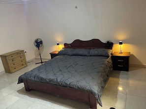 A bedroom with a bed, two bedside tables with lamps, a wooden dresser, and a standing fan. - From home to home accommodation with the space you need to make a perfect stay. (Freetown)
