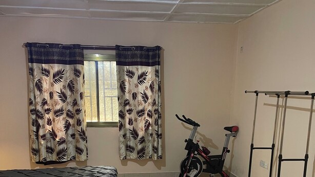 Une chambre avec un lit, une fenêtre ornée de rideaux à motifs, un vélo d’appartement et un porte-objets fixé au mur.