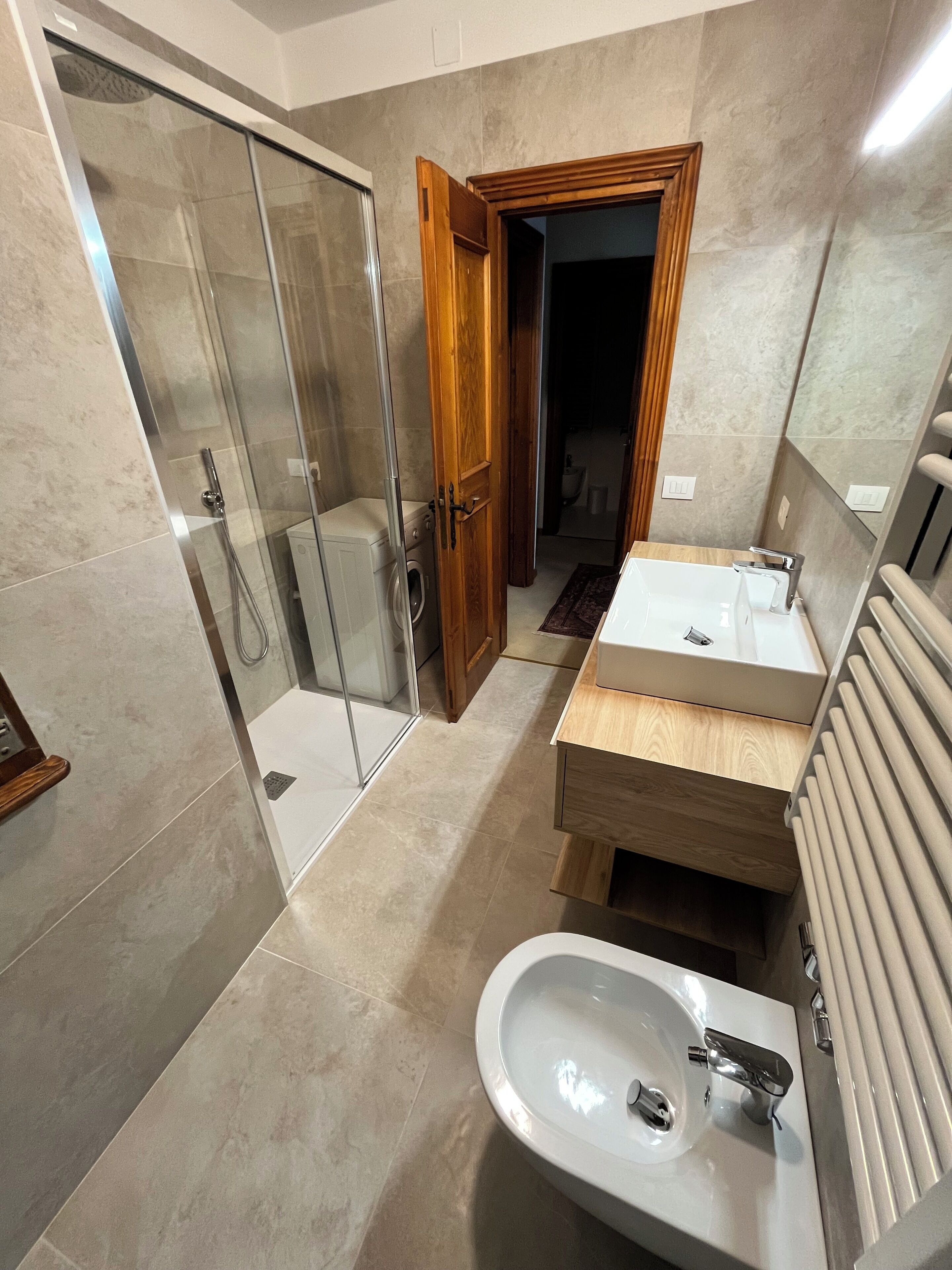 A modern bathroom with a glass shower enclosure, a wooden door, a white sink, and a built-in radiator.