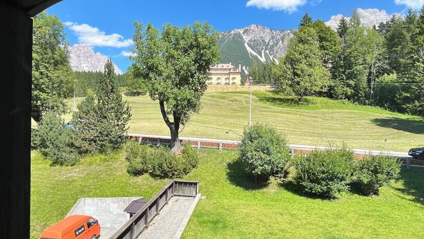 A view from inside a building looking out to a grassy area with trees, a building, and mountains in the background.