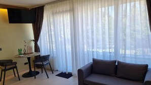 Habitación de hotel con sofá, una pequeña mesa con sillas, televisor montado en la pared y ventanas amplias con cortinas traslúcidas.