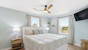 A bedroom with a bed, nightstand, lamp, ceiling fan, TV, windows, curtains, and a view of the outside. - New Listing, Flex Stays, Dog Friendly, Close to Beach and Pier, KD0105 (Kill Devil Hills)
