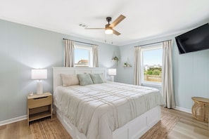 A bedroom with a bed, nightstand, lamp, ceiling fan, TV, windows, curtains, and a view of the outside.