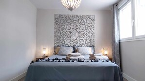 Un dormitorio con una cama grande, dos lámparas de noche, un panel decorativo en la pared y una ventana con cortinas.