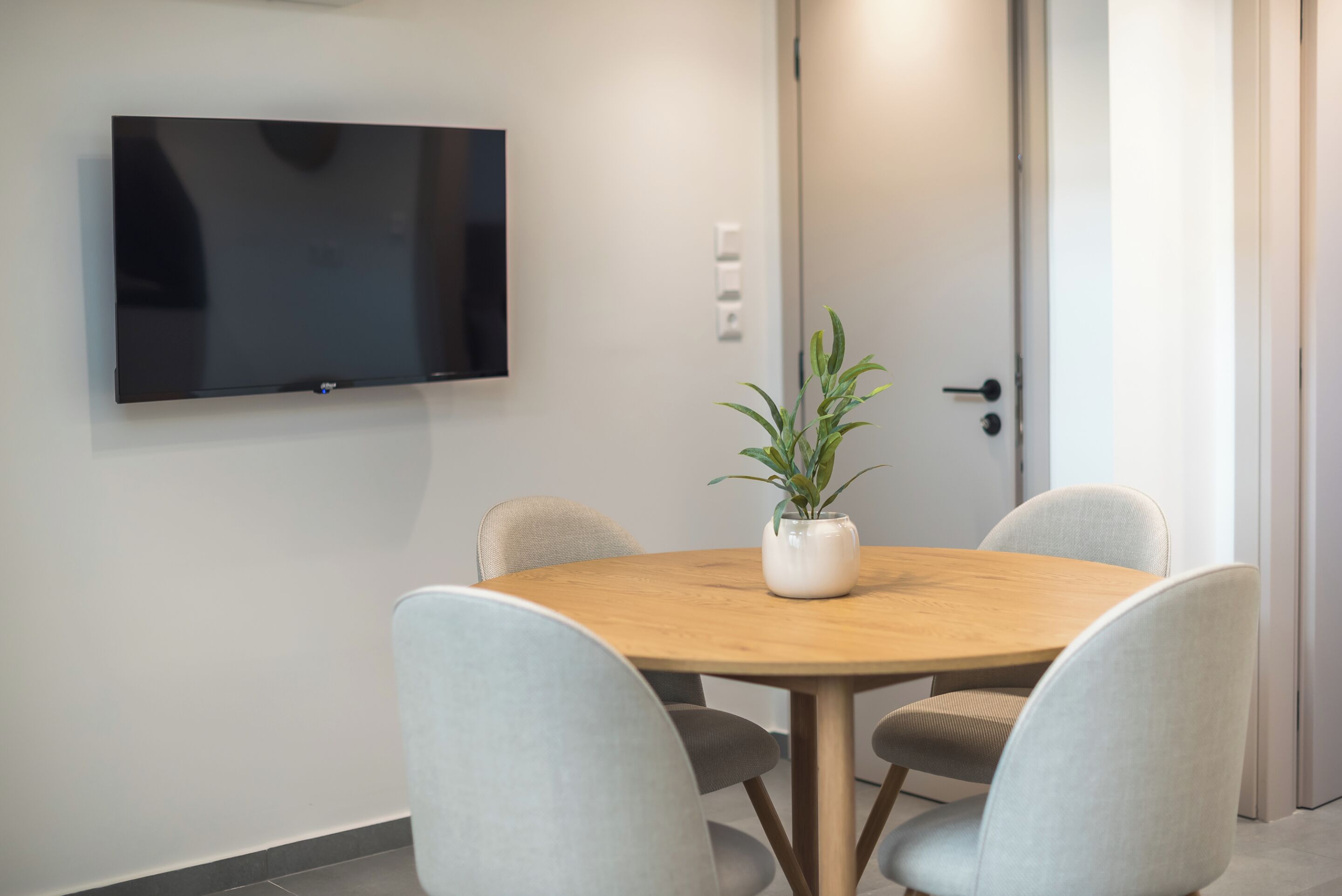 A conference room with a round wooden table, four grey chairs, a potted plant, and a flat-screen TV mounted on the wall.