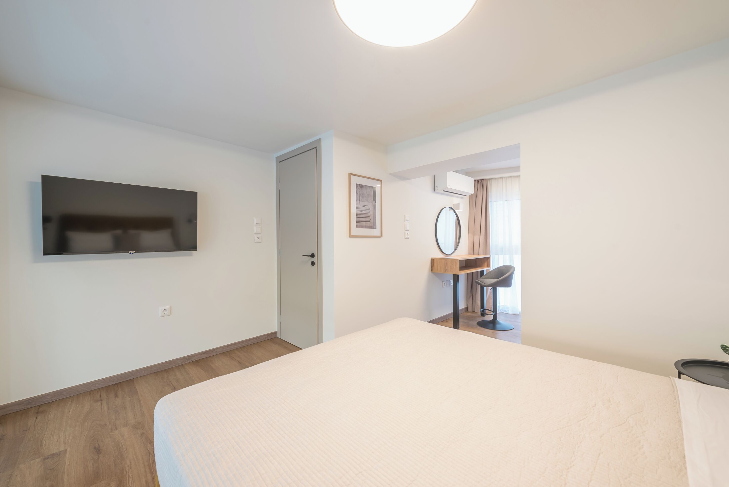 A bedroom with a bed, a TV mounted on the wall, a door, a mirror, and a small desk with a chair.
