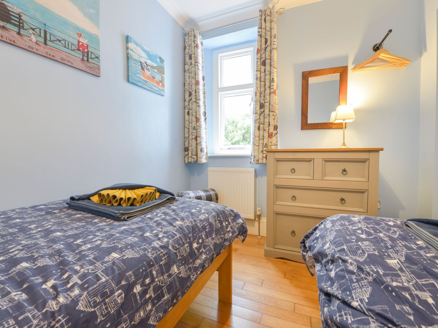 Quarto com duas camas, cômoda, abajur e janela com cortinas.