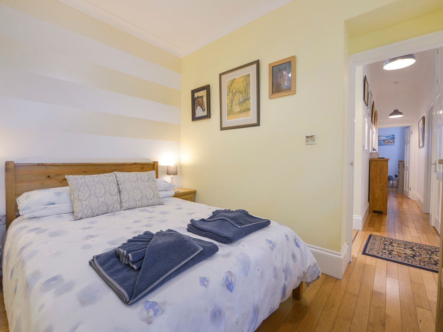 A bedroom with a bed, pillows, a nightstand, and a lamp. There are framed pictures on the wall and a hallway leading to another room.