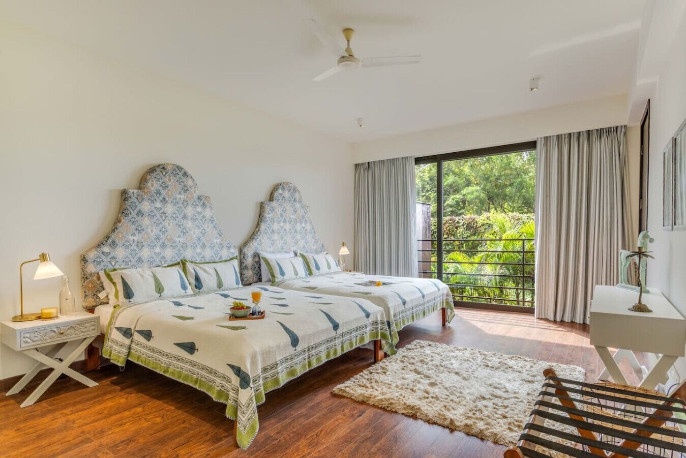 Una camera da letto con due letti, un balcone vista verde, un ventilatore a soffitto e un tavolino con una lampada.