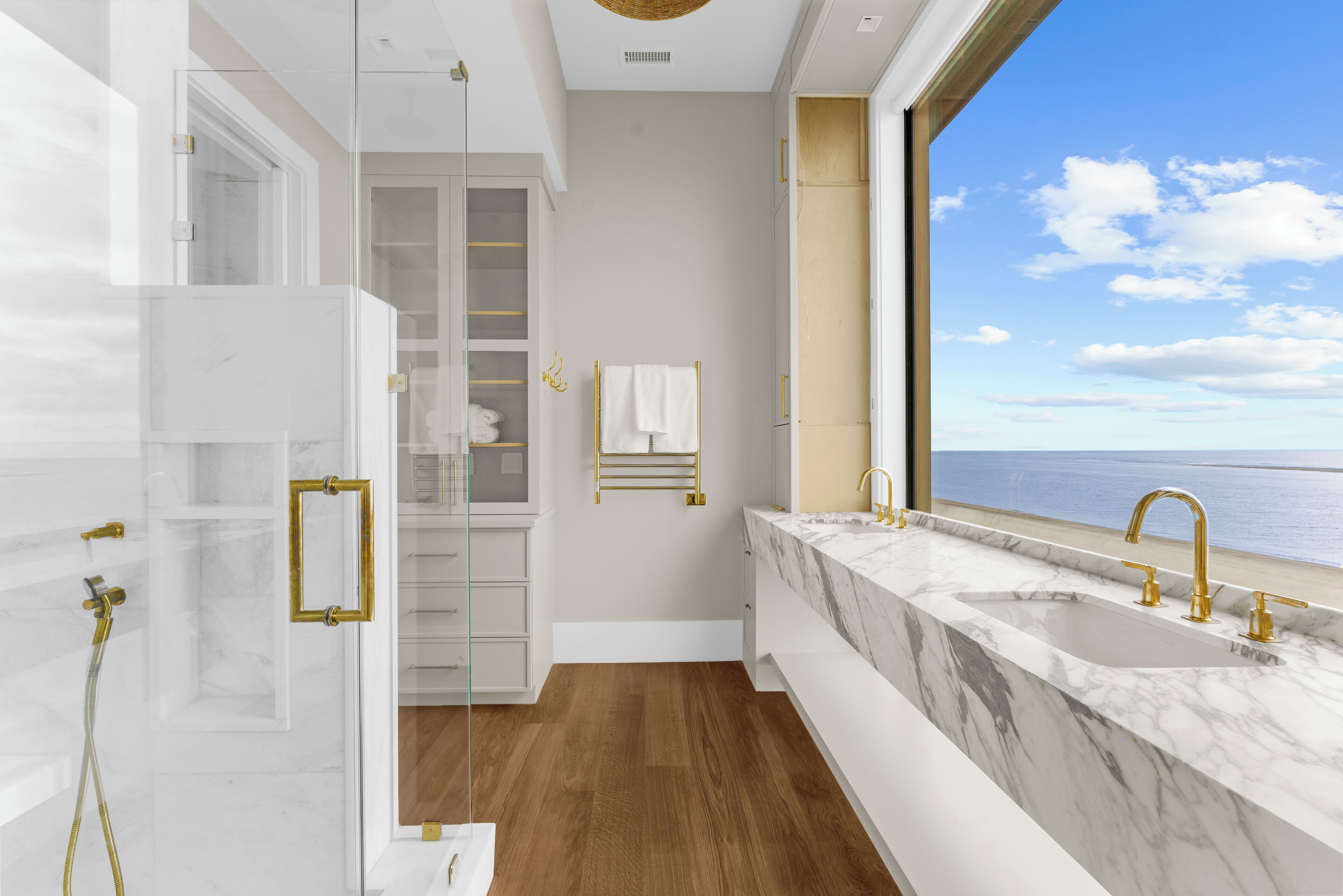 Un baño moderno con un ventanal grande, cabina de ducha, lavamanos de mármol y suelo de madera.