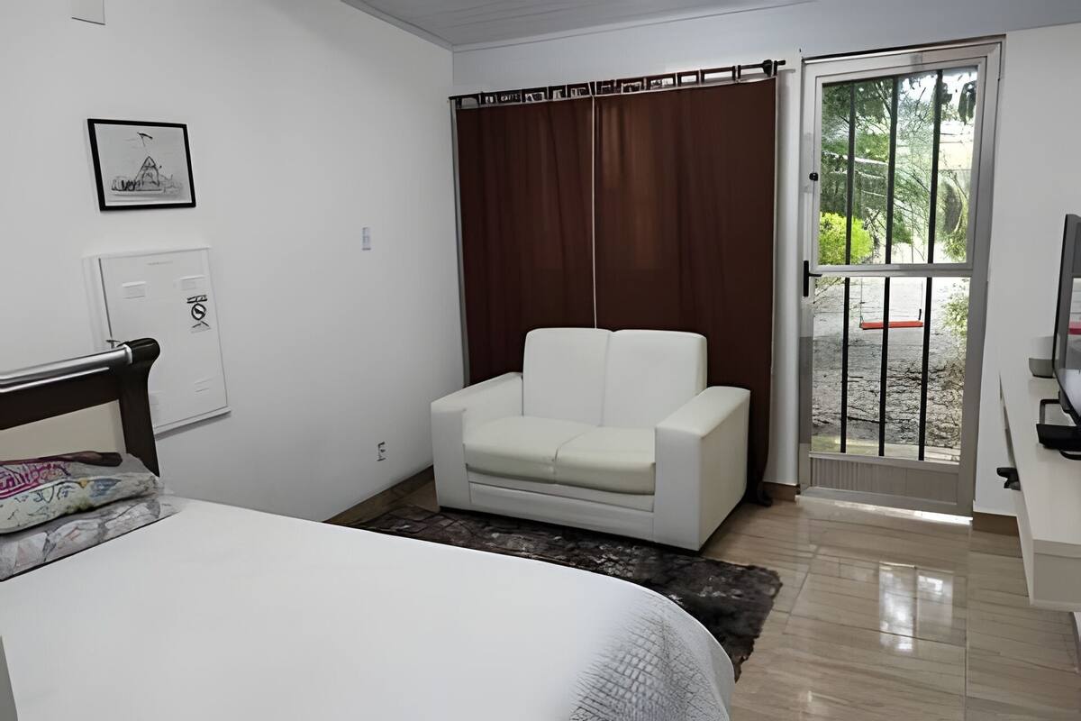 Une pièce comprenant un lit, un fauteuil blanc, un petit tapis, une porte donnant sur une vue agréable et un interrupteur fixé au mur.