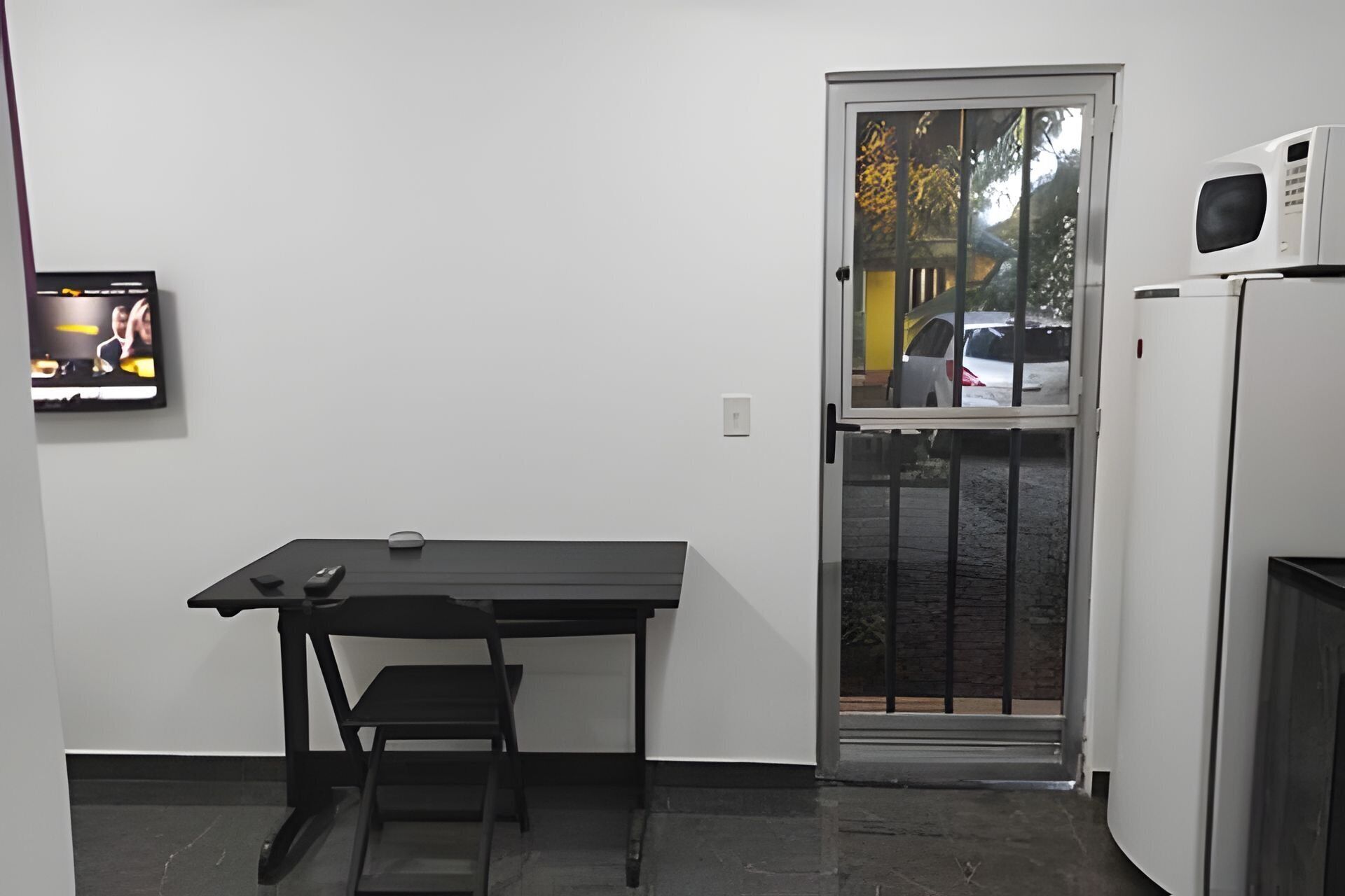 Habitación pequeña con escritorio, silla, televisión y puerta que da a vista de coches estacionados afuera.