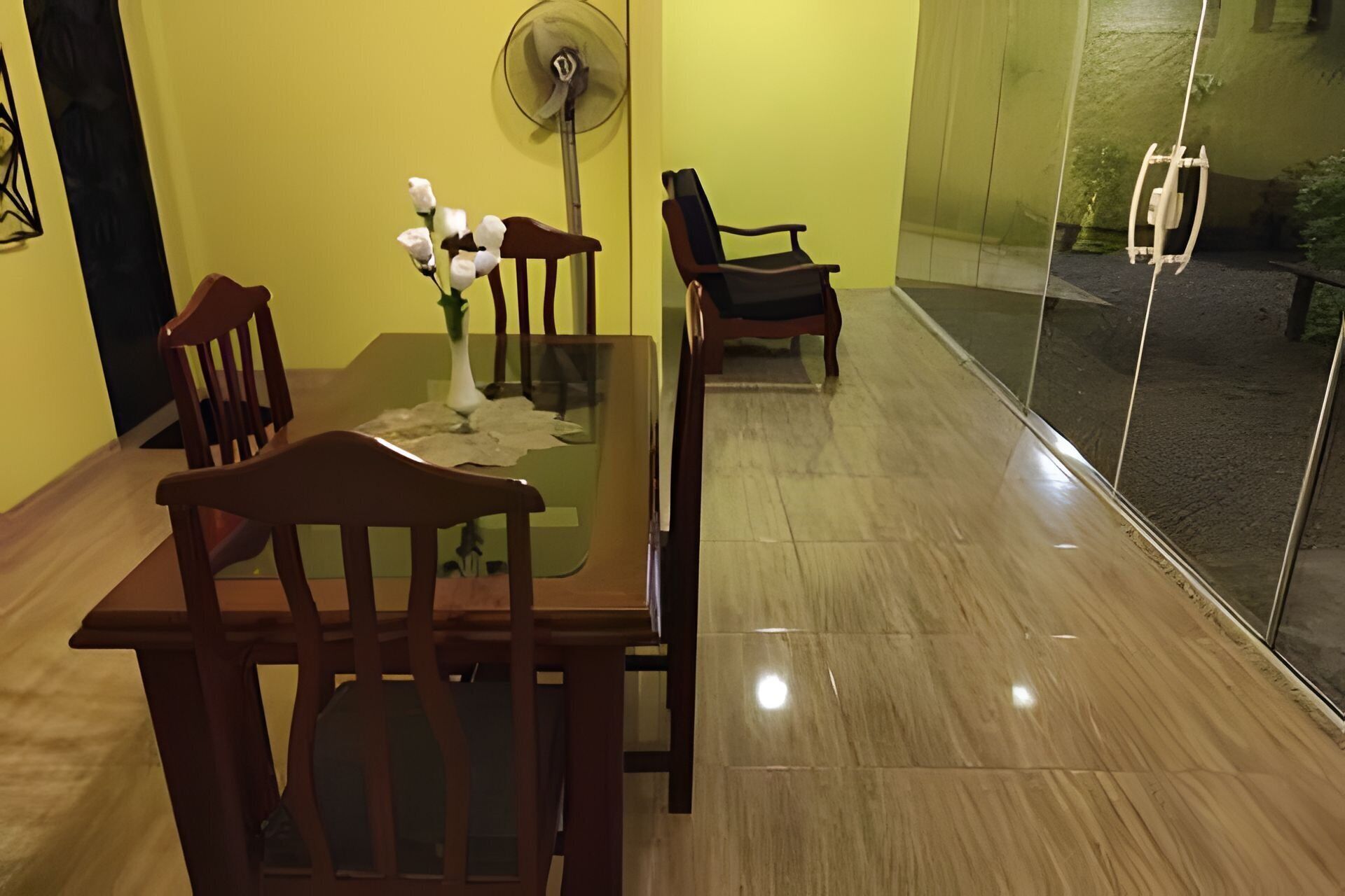 A dining area with a glass table, wooden chairs, a vase with flowers, and a standing fan.