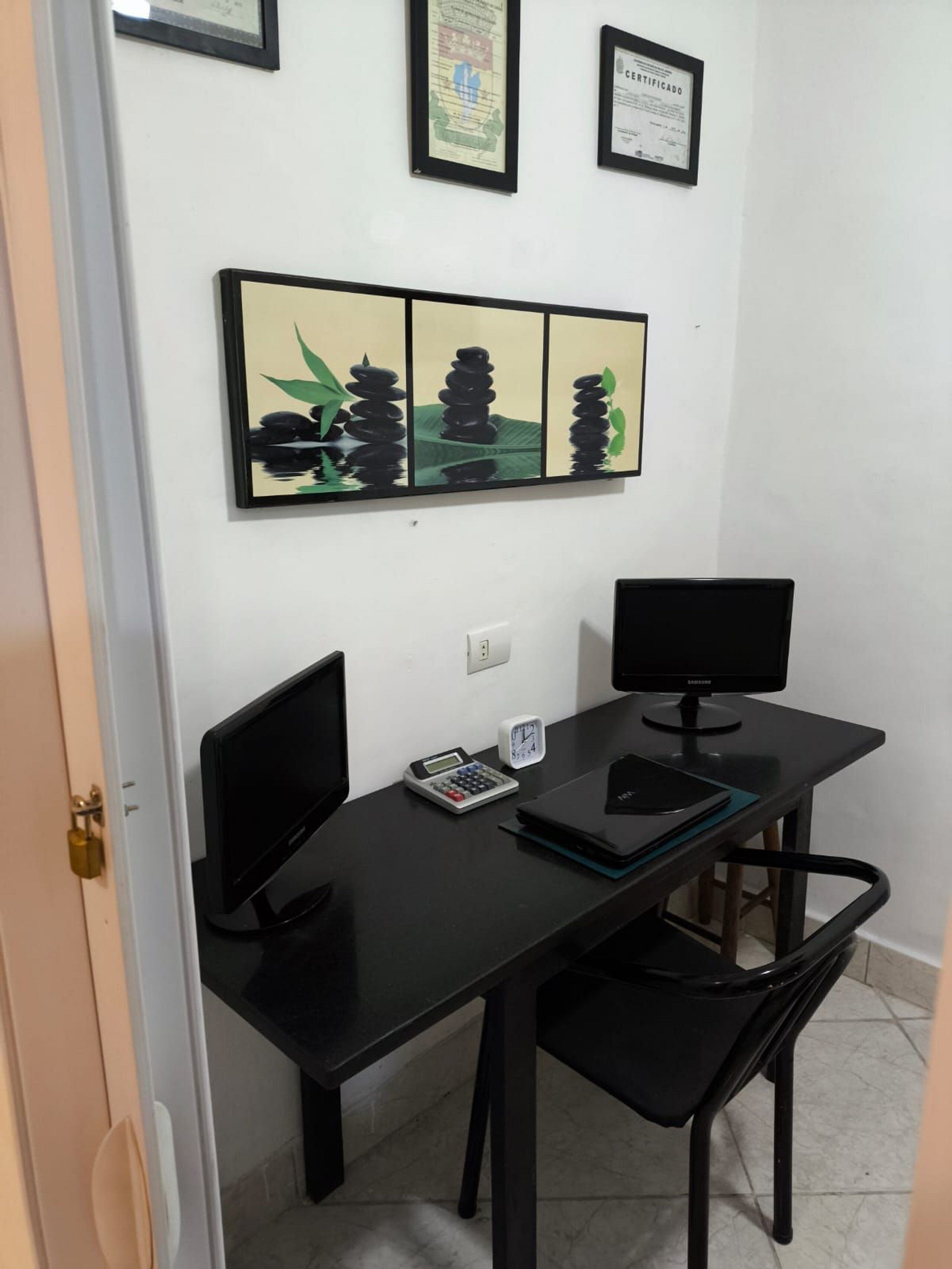 A room with a black desk, two monitors, a calculator, and a laptop. There are certificates on the wall.