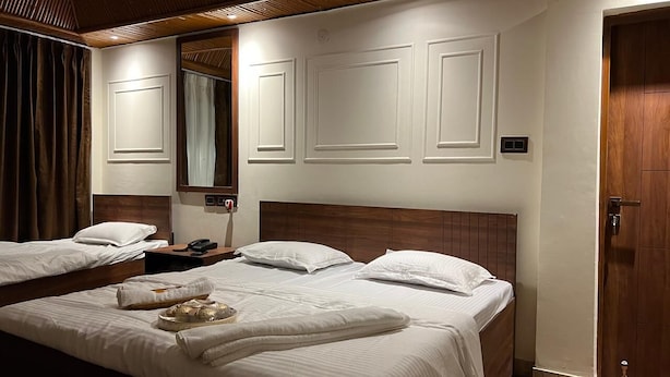 A hotel room with two beds, a wooden headboard, a bedside table with a lamp, and a large mirror on the wall. - Ambika Resort (Rishikesh)