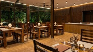 A restaurant with wooden tables and chairs, ceiling fans, and a view of greenery through large windows. - Ambika Villa - A Unit Of D-Musk Hospitality (Rishikesh)
