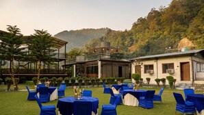 An outdoor seating area with tables and chairs, a covered structure, and a building with a white facade. - Ambika Resort (Rishikesh)