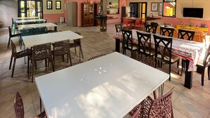 Un comedor interior amplio con techos altos y vigas a la vista, con varias mesas y sillas, una zona de cocina y un televisor.