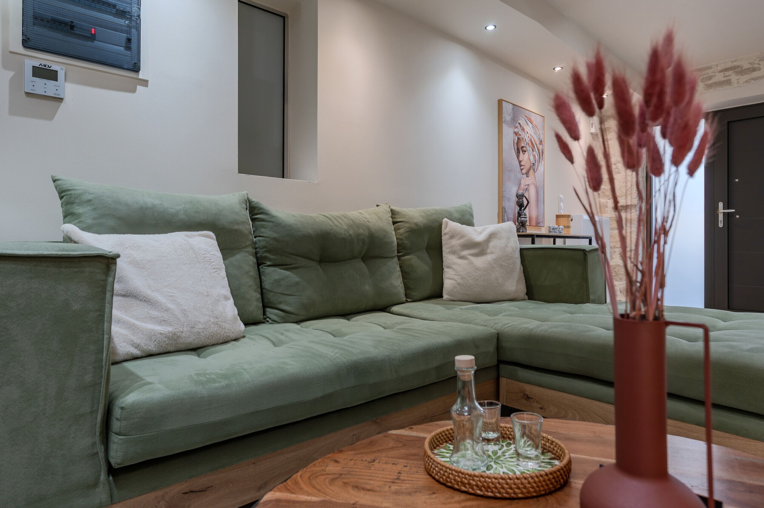 A green sofa with white and green cushions, a wooden coffee table with a glass and bottle, and a vase with dried plants.
