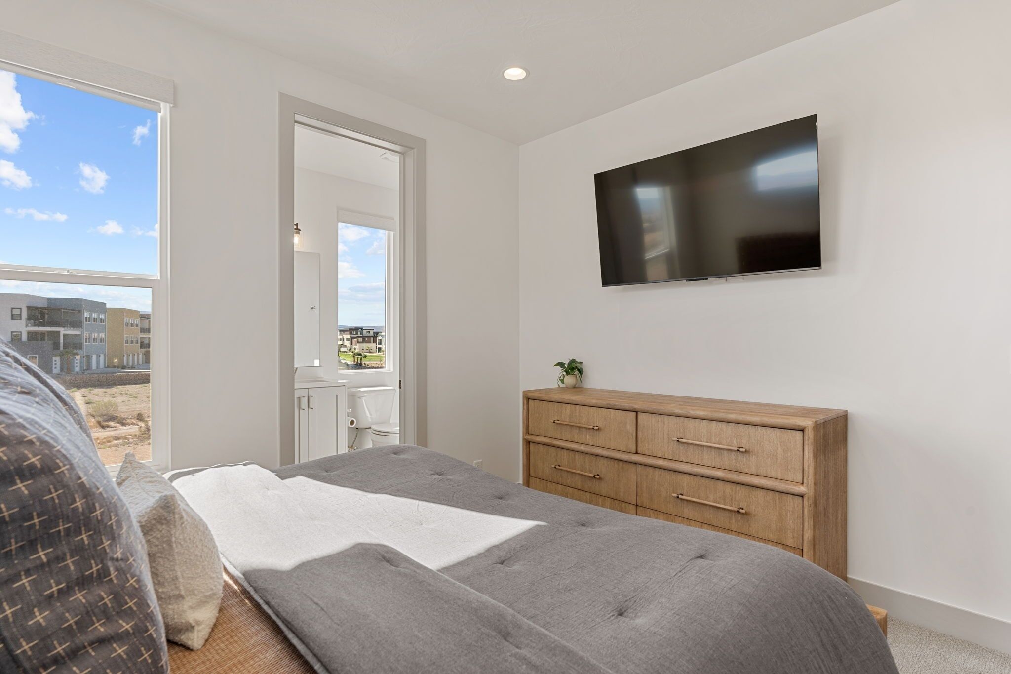 A bedroom with a bed, a wooden dresser, a TV on the wall, and a window with a view of buildings.