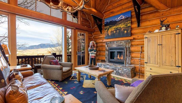 A log cabin interior with a stone fireplace, wooden furniture, and a large window overlooking a mountainous landscape. - Summit Cabin 3 Bedroom Home (Telluride)