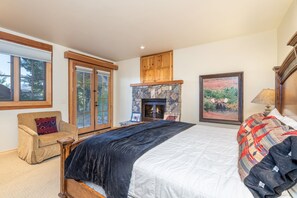 A bedroom with a large bed, a stone fireplace, wooden cabinets, and a window with a view of trees. - Summit Cabin 3 Bedroom Home (Telluride)