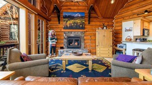 A log cabin interior with a stone fireplace, wooden furniture, and a view of mountains through the window. - Summit Cabin 3 Bedroom Home (Telluride)