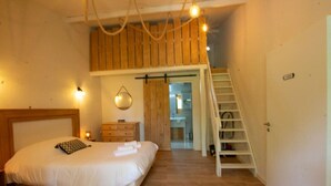 Ein Schlafzimmer mit Bett, Holz-Kopfstück, Nachttisch, Lampe und einer Wendeltreppe zu einem Schlafbereich im oberen Geschoss.